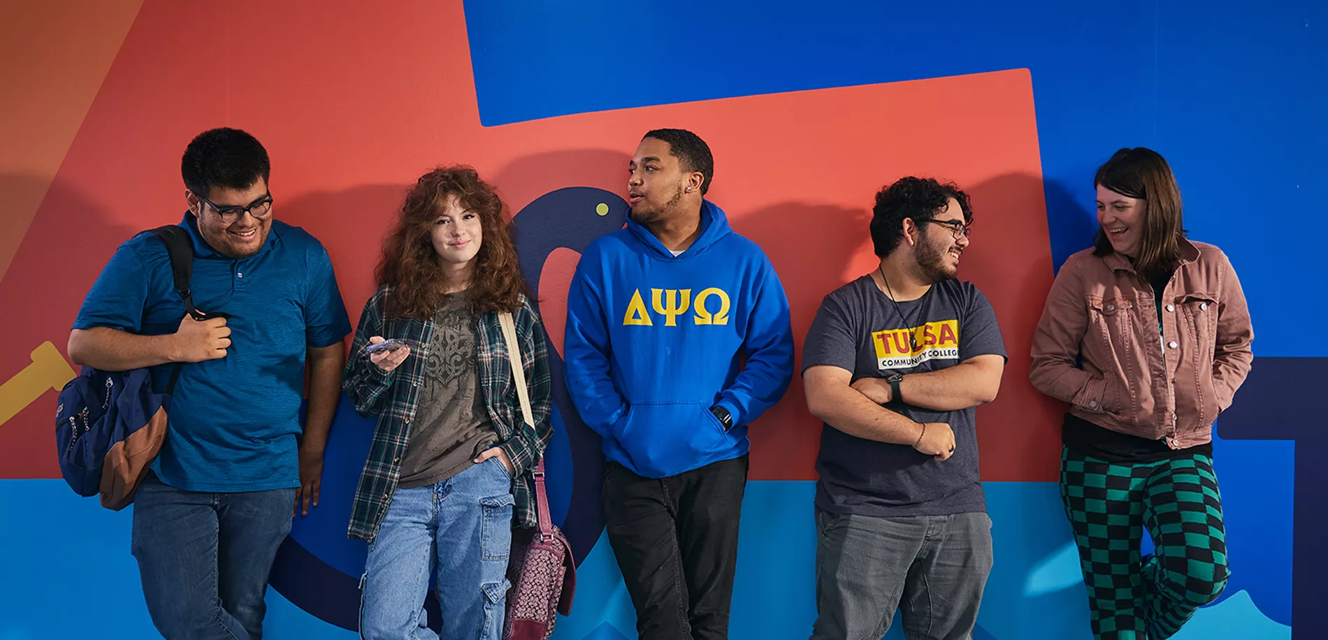 A group of five TCC students stands against a colorful wall. They are casually dressed, with some holding backpacks.