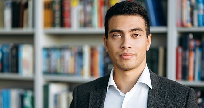 Hispanic Man in a blue collard shirt and blazer. A full bookshelf fills the background.