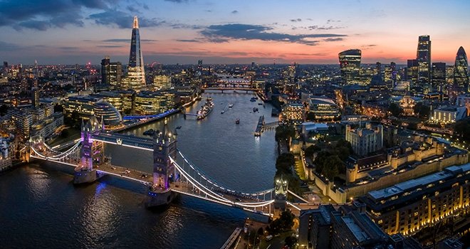 The sun sets behind a London skyline. The Tower Bridge and the River Thames are in the foreground with the city stretching into the horizon.