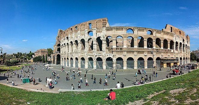 The Roman Colosseum towers over the crowds of visitors that mill around its courtyard under a clear blue sky.