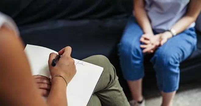 View over the should of a counselor notebook in hand. Patient, seen from the shoulders down, sits on a couch, hands folded.