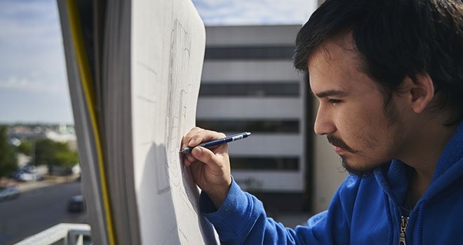 man using a pencil to draw on a large canvas on an easel in an urban setting.