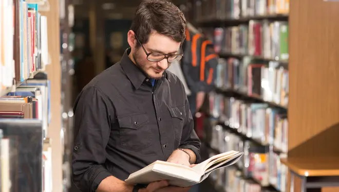 A man is standing and reading a book with library shelves full of book behind him.