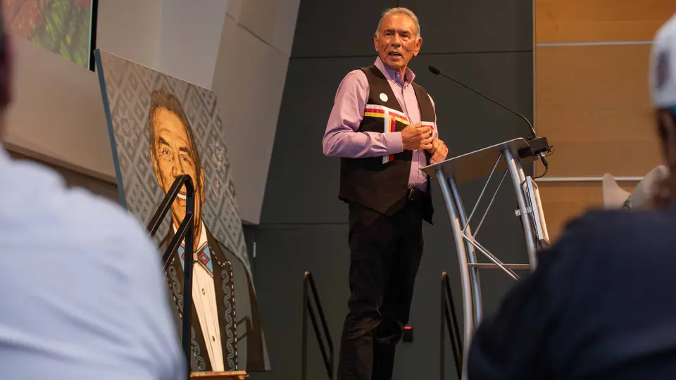 Wes Studi stands a podium and speaks to a crowd of people.